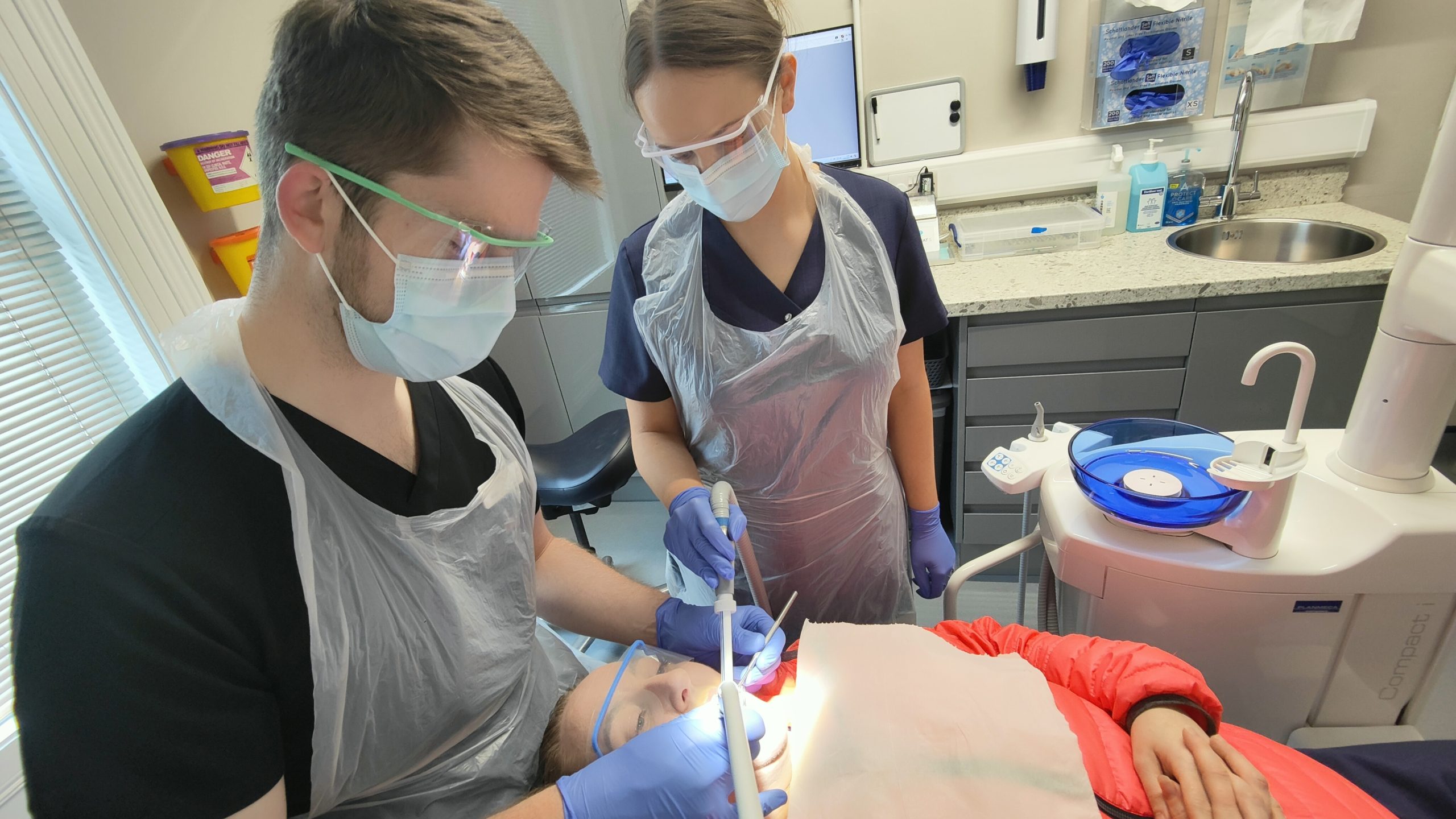 Two dental professionals performing a procedure on a patient in a dental chair. The patient wears a red jacket and protective glasses, while the professionals wear gloves, masks, and aprons. Dental tools and equipment are visible in the background.