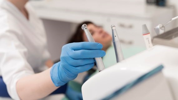 A dental professional in a white coat, blue gloves, and a face mask holding a dental tool, with a blurred patient seated in a dental chair and dental equipment visible in the background.