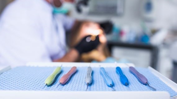 Tray of five dental tools with colored handles (green, pink, gray, blue, and purple) in the foreground, with a blurred view of a dentist treating a patient in the background.