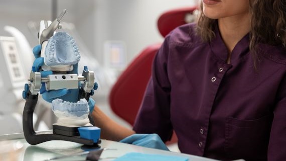 Dental professional in a purple uniform and gloves holding a dental model with an articulator, used to simulate jaw movement and tooth alignment, in a clinical or lab setting.