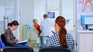 Dental clinic waiting area with two people seated, one writing on a clipboard and the other looking toward the treatment area. In the background, a dentist is attending to a patient in a dental chair, with visible equipment, a computer monitor, and an anatomical poster of teeth.