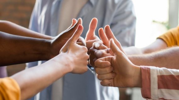 A group of people standing in a circle with their hands extended towards the center, thumbs up, symbolizing unity and teamwork. The image is blurred in the center to focus on the hands and thumbs.