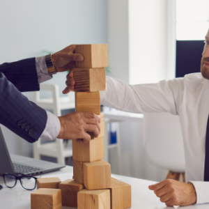 Two individuals in a professional setting are stacking wooden blocks on a desk. One wears a dark suit, the other a white shirt with a black tie. A laptop and eyeglasses are also on the desk. Their faces are blurred for privacy.