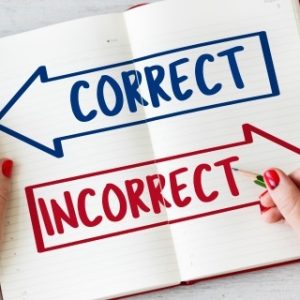 An open notebook displays two large arrows: a blue arrow labeled 'CORRECT' on the left and a red arrow labeled 'INCORRECT' on the right. One person with red nail polish holds the notebook, while another points at the 'INCORRECT' arrow with a green pencil.