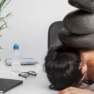Person sitting at a desk with head resting on the surface, four large stones stacked on their head. Desk contains a potted plant, pencils in a cup, water bottle, glasses, and notebooks.