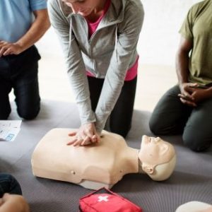 A group of people gathered around a CPR training session, with one person performing chest compressions on a CPR manikin. Others are observing and possibly taking notes. A red first aid kit is visible in the foreground.