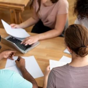 Four people sit around a wooden table in a collaborative setting, holding papers and working together. One person is using a laptop, suggesting a study or teamwork environment.